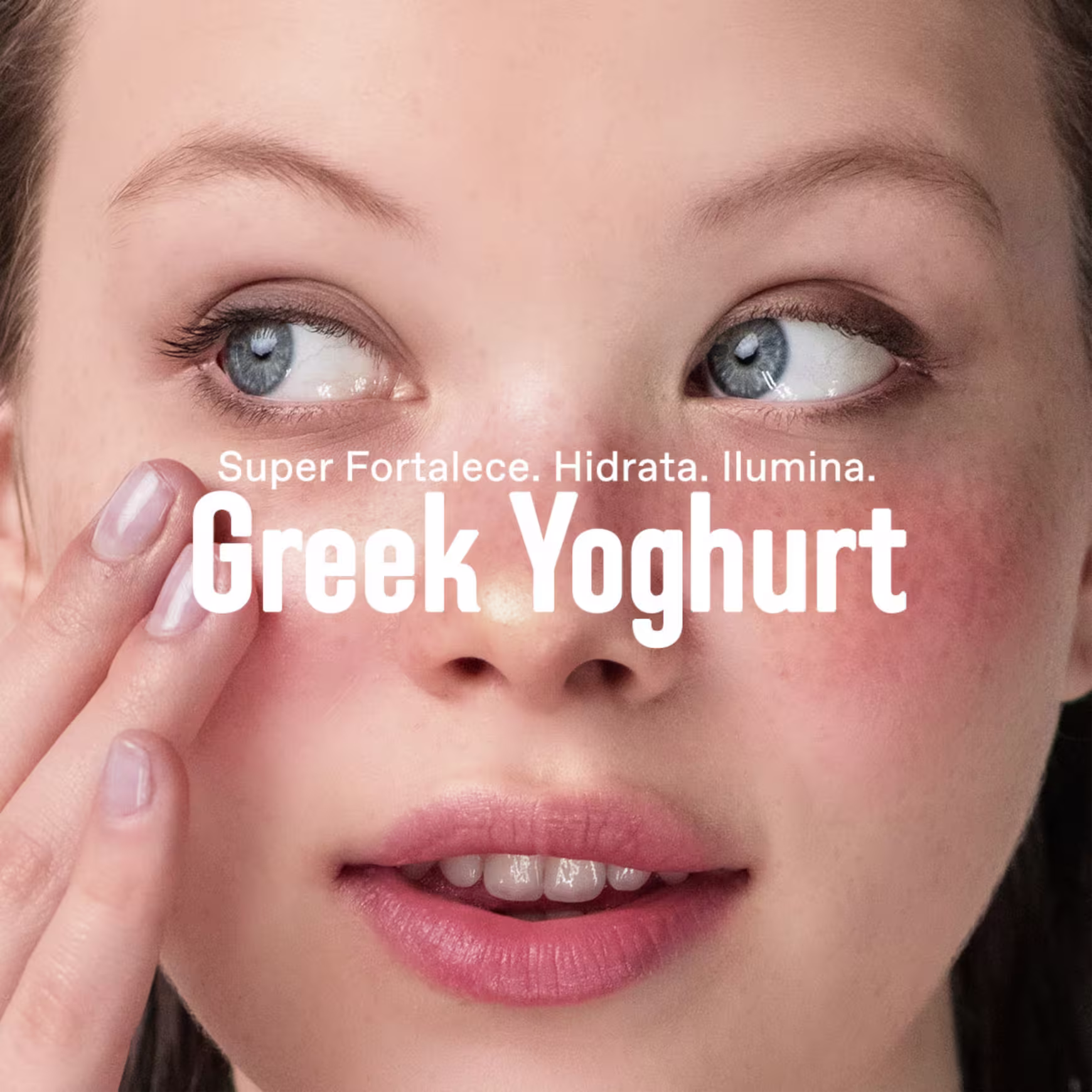 A imagem mostra um close-up do rosto de uma mulher, com seus olhos azuis e lábios rosados em destaque. O texto "Super Fortalece. Hidrata. Ilumina. Greek Yoghurt" está sobreposto na imagem, sugerindo que o iogurte grego é um ingrediente chave.