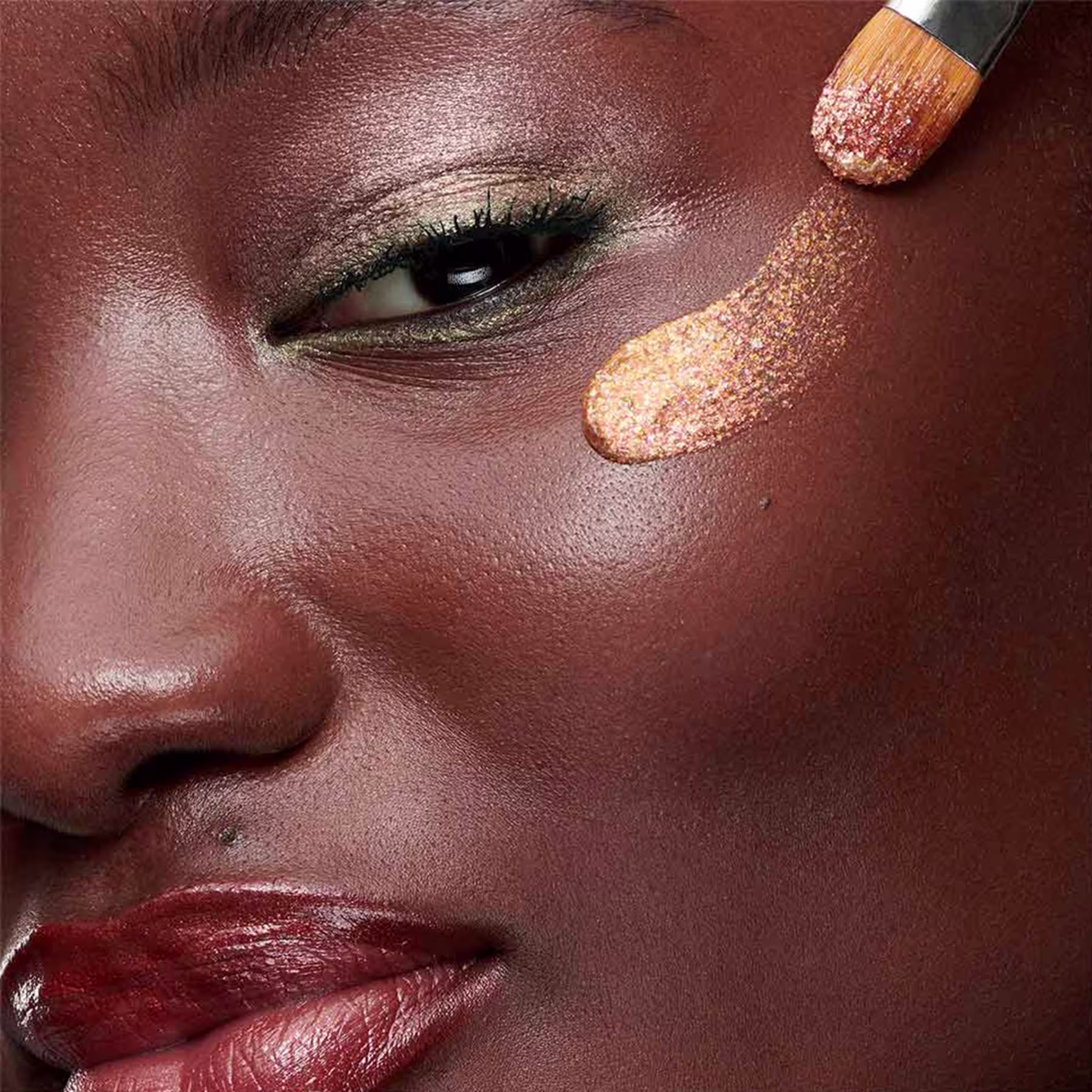 A imagem mostra a modelo com um pincel passando o glitter no rosto.