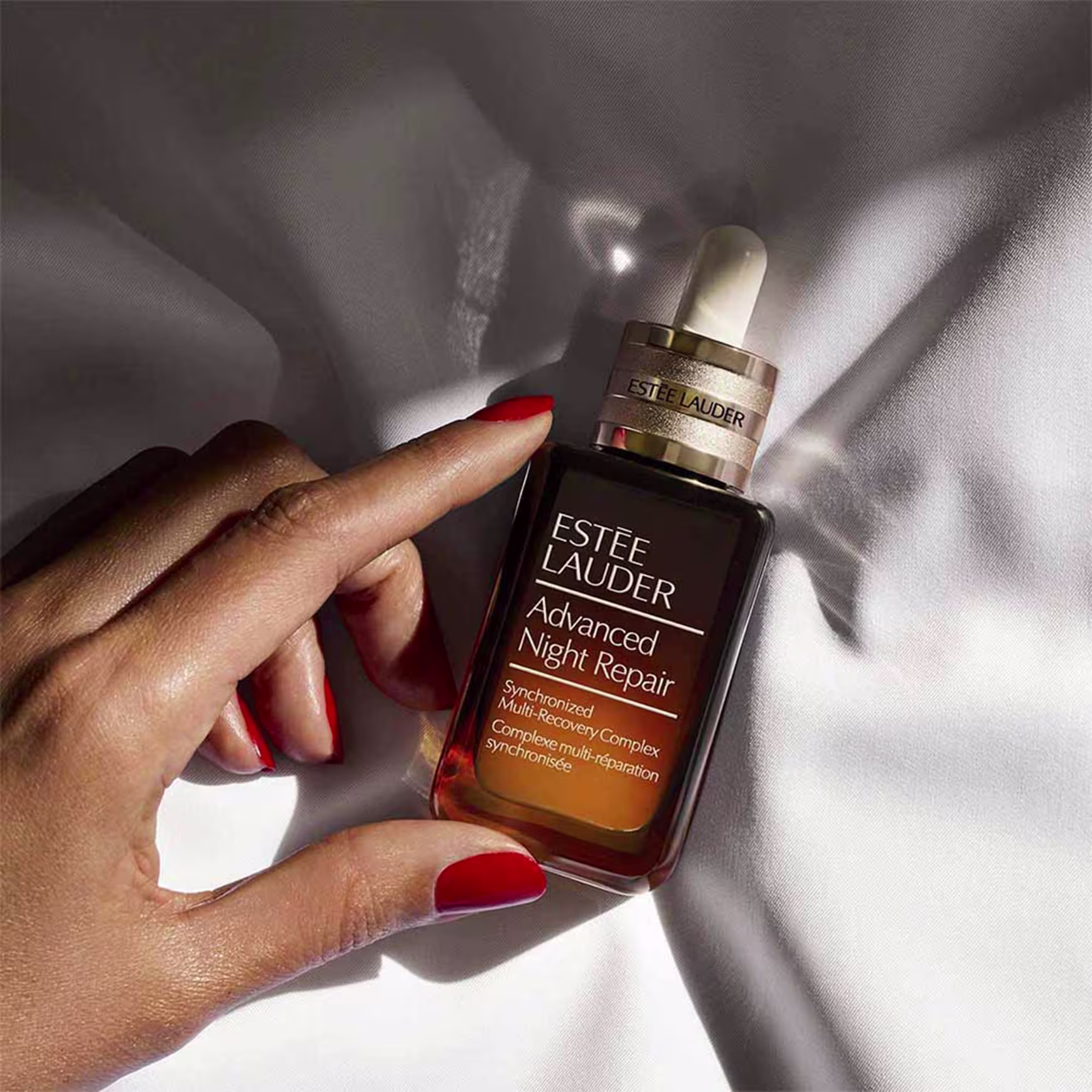 Produto de tratamento noturno para a pele da marca Estée Lauder.  Rótulo com o nome "Advanced Night Repair" e descrição "Synchronized Multi-Recovery Complex" em letras brancas. Uma mão feminina segura o frasco, com unhas pintadas de vermelho.
