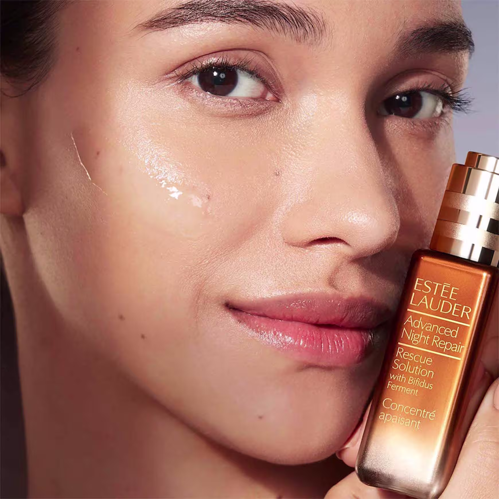 Sérum facial da linha Advanced Night Repair Estée Lauder. O rótulo exibe o nome do produto "Advanced Night Repair Rescue Solution with Bifidus Ferment" em letras douradas sobre um fundo laranja. O frasco está sendo segurado próximo ao rosto de uma pessoa.