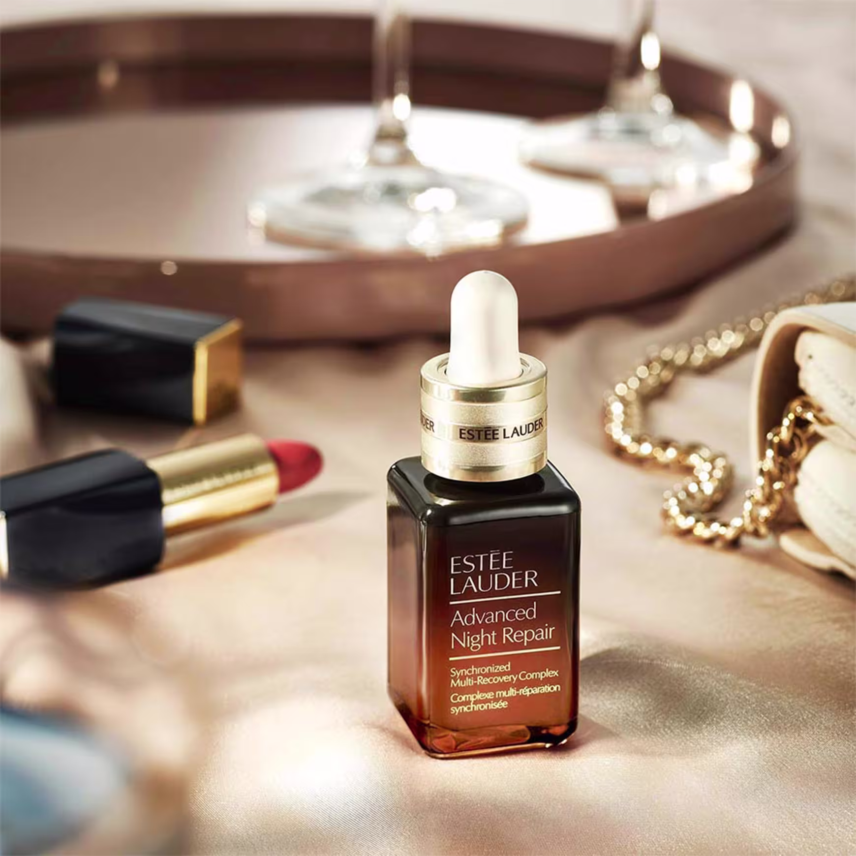 Frasco com rótulo "Estée Lauder Advanced Night Repair". Conta-gotas branco na parte superior. Tubos de batom Estée Lauder em vermelho e dourado. Colar de pérolas disperso. Luzes pendentes borradas ao fundo, criando uma atmosfera suave e elegante.