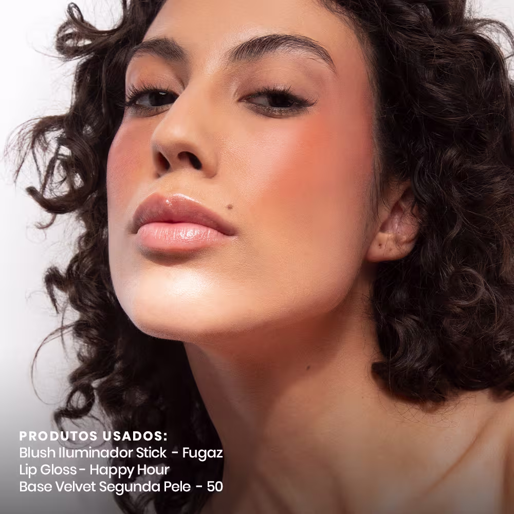 A imagem mostra uma modelo maquiada com blush iluminador em bastão Fugaz, lip gloss Happy Hour e base Velvet Segunda Pele