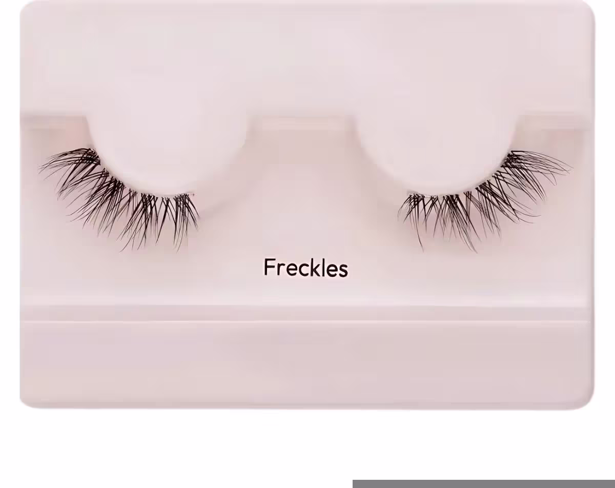 A imagem mostra os cílios postiços em um estojo plástico claro. O estojo tem uma cor nude claro e exibe um par de cílios postiços pretos.