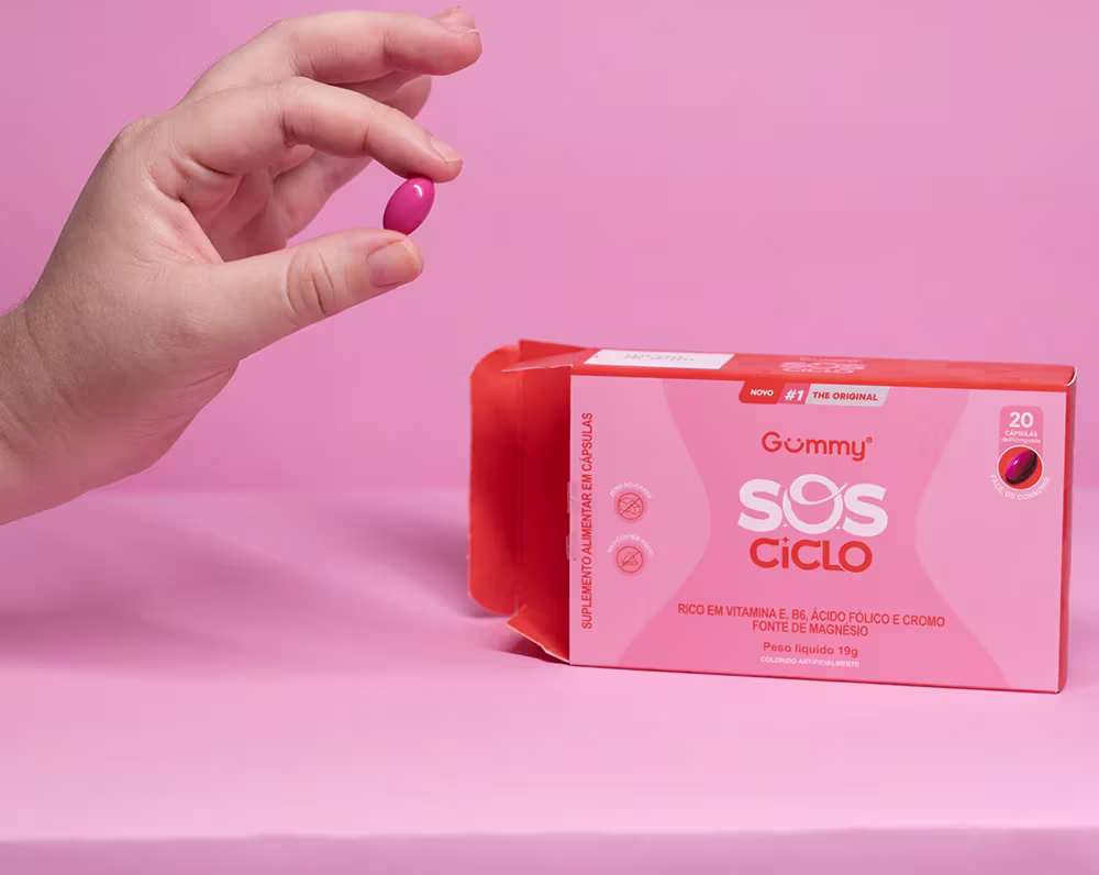 Caixa de suplemento vitamínico Gummy SOS Ciclo. A embalagem tem texto em português "Rico em vitamina E, B6, ácido fólico e cromo - Fonte de magnésio". Uma mão feminina segura um comprimido vermelho próximo à caixa, sobre um fundo rosa claro.