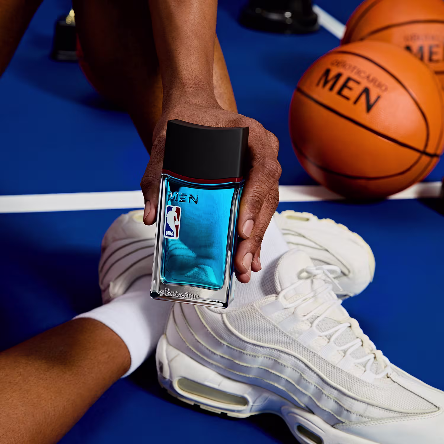 O frasco possui um design retangular com uma tampa preta e a logomarca da NBA impressa em azul.Possui uma coloração azul predominante. O produto está posicionado junto a uma bola de basquete laranja, sugerindo uma associação com o esporte.