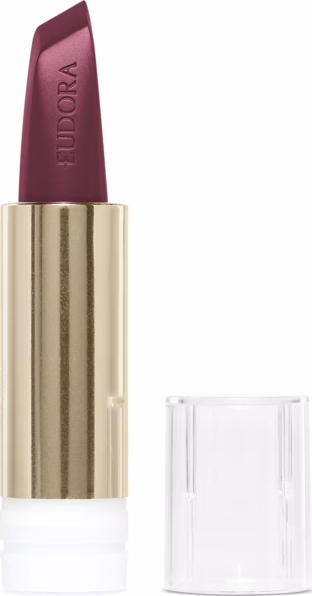 A imagem mostra o batom Eudora na sua case refil. A case está aberta, é dourada e a tampa é transparente. O Batom tem formato de bala de revólver e está todo à mostra, sua cor é de tom vinho, elegante, escuro e rico. A palavra Eudora está gravada no batom