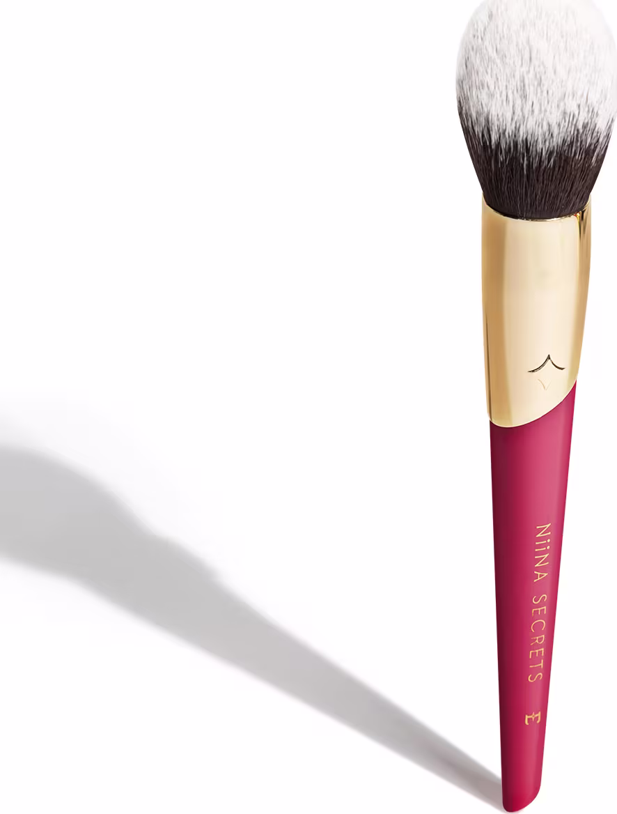 Pincel de maquiagem com cabo rosa e dourado. Parte rosa do cabo tem Niina Secrets impresso em dourado. A parte superior é dourada. O pincel tem cerdas brancas e marrons, formando uma cabeça arredondada e densa.