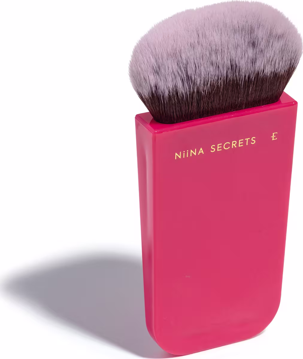 O produto apresentado é um pincel de maquiagem da marca Niina Secrets. O pincel possui cerdas macias e uma base em um tom rosa vibrante, com o nome da marca impresso em letras douradas. Em outro ângulo.