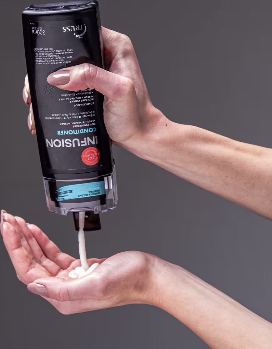 A imagem mostra uma mão segurando um frasco de condicionador da marca Truss, linha Infusion. Também é possível ver o seu conteúdo, que é cremoso e branco, com 300ml.