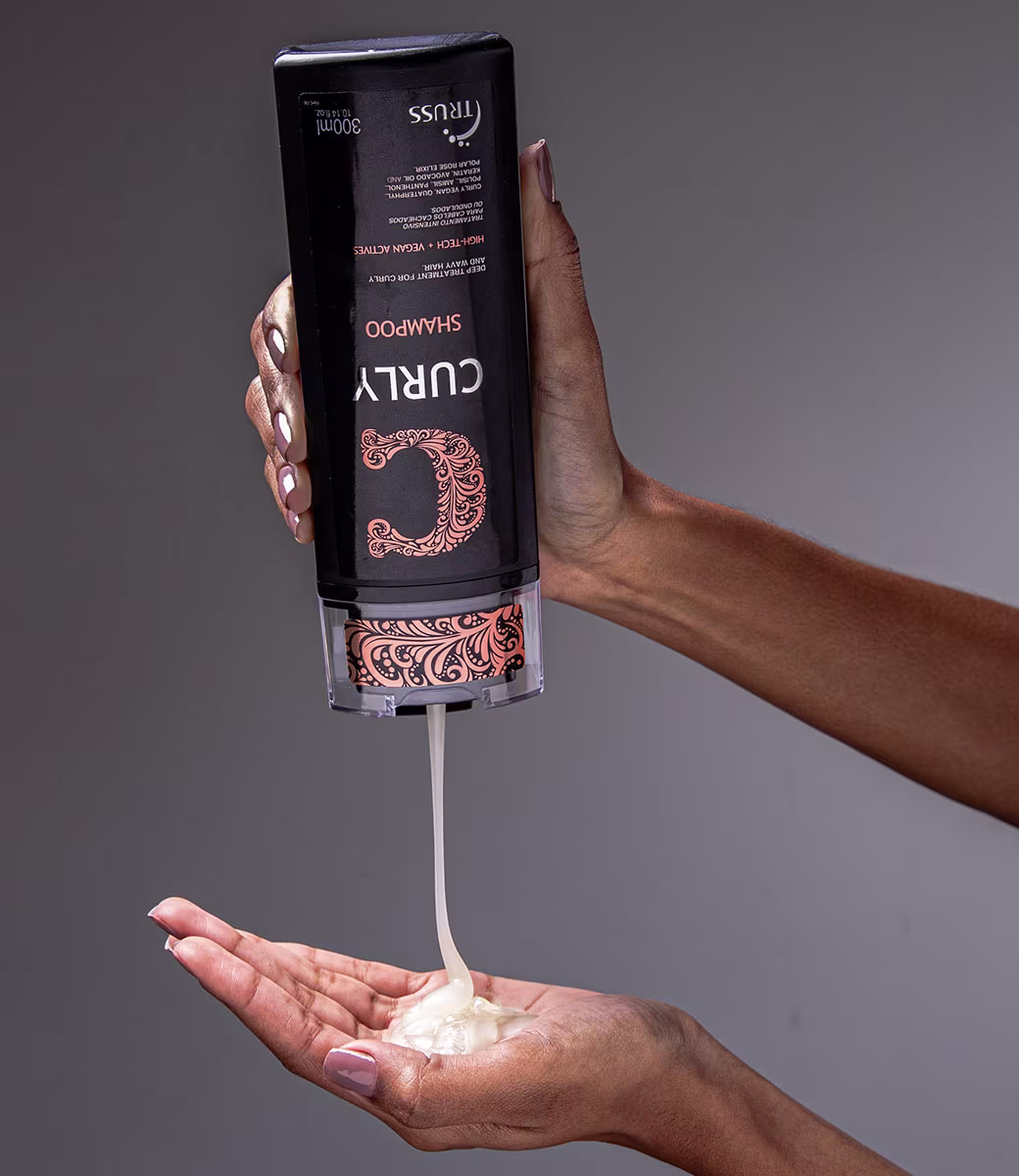 A imagem mostra o Shampoo TRUSS Curly. O líquido está sendo despejado do frasco em uma mão feminina, mostrando a espessura do shampoo.