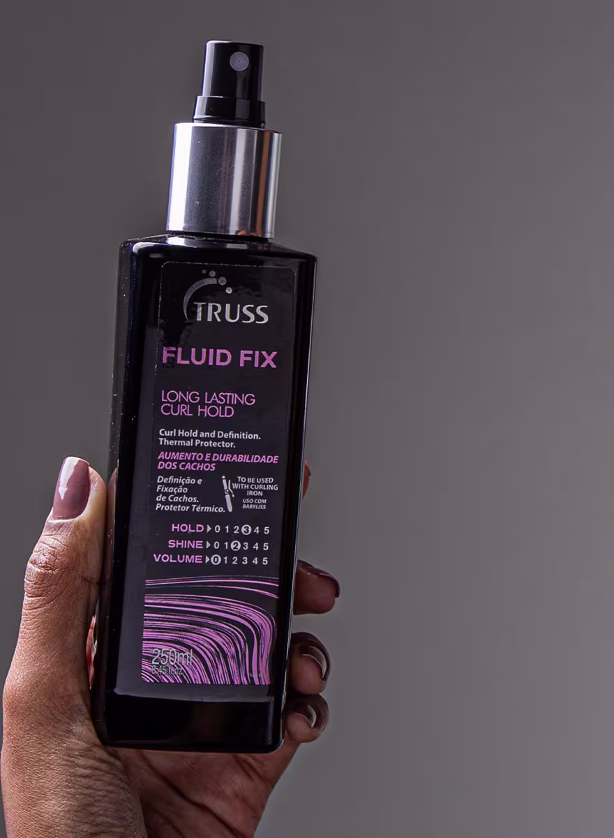 Com fundo cinza, a imagem mostra uma mão feminina segurando o ativador de cachos Fluid Fix, da marca TRUSS: um frasco preto com pulverizador metálico prateado e detalhes em roxo. Destaca o benefícios de aumento e durabilidade dos cachos.