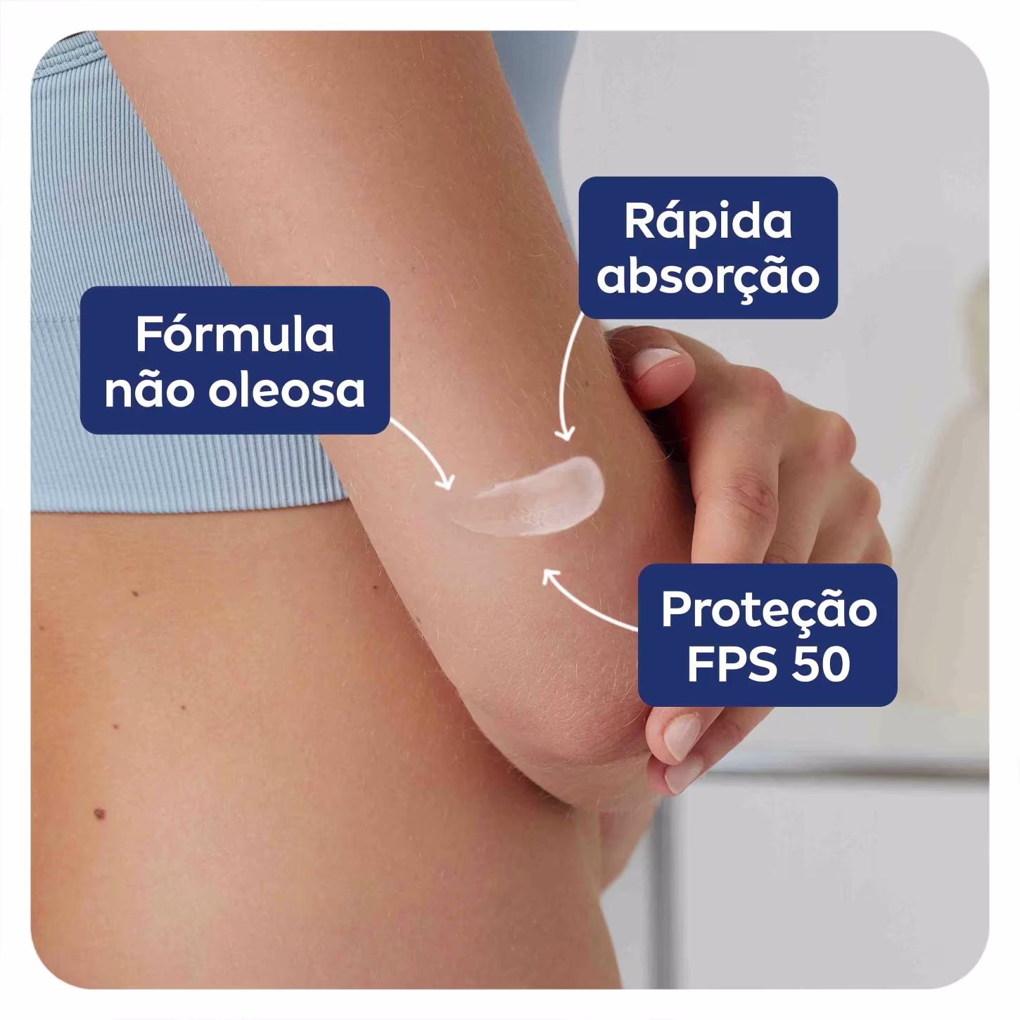 A imagem mostra uma mão aplicando o produto nos braços perto do cotovelo. As cores predominantes são tons de pele humana, azul dos rótulos textuais e branco do produto aplicado.