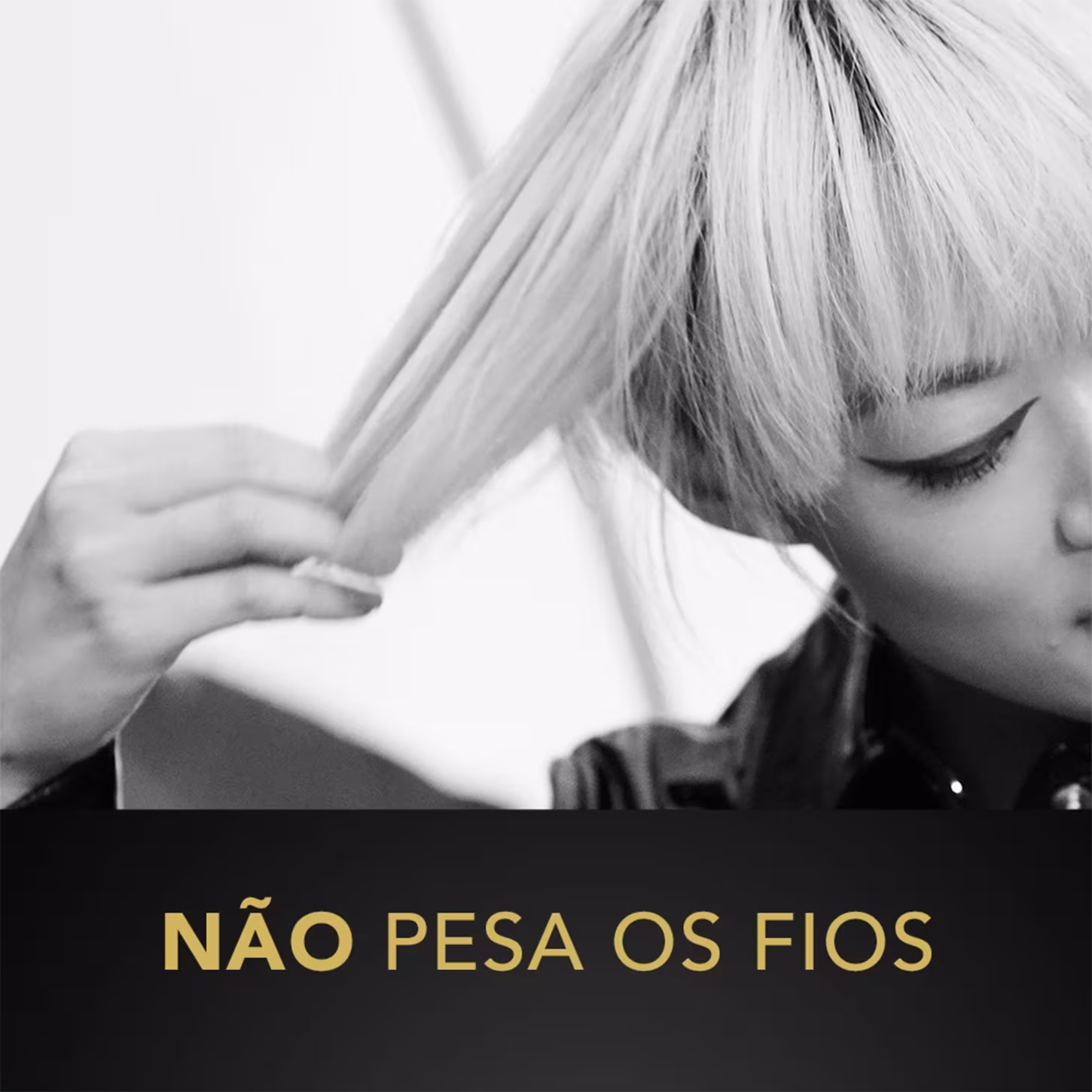 A imagem mostra uma pessoa com cabelos loiros cobrindo parcialmente o rosto. Apenas um dos olhos é visível, com maquiagem escura destacando os cílios. A mão da pessoa segura uma mecha do cabelo loiro na frente do rosto.