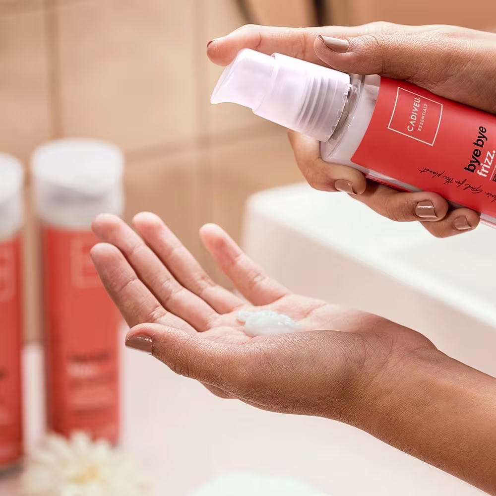 Uma mão está derramando o leave-in na outra. Ao fundo os demais produtos da linha.