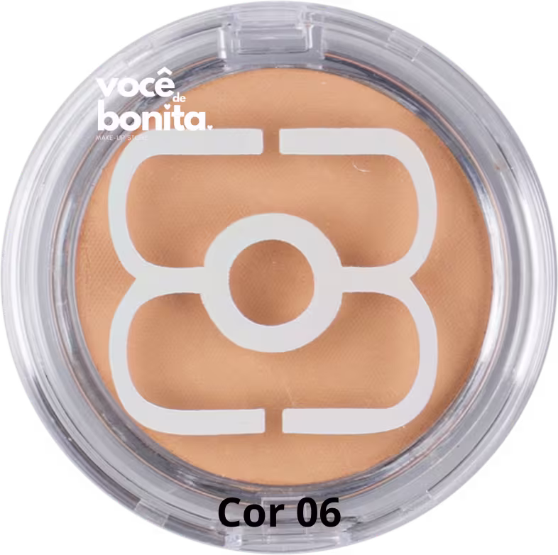 Pó compacto facial em embalagem redonda transparente de plástico. A cor do produto é um tom de bege claro. No centro da embalagem há um símbolo estilizado em branco. O rótulo na parte inferior exibe o texto "você bonita" e "Cor 06".