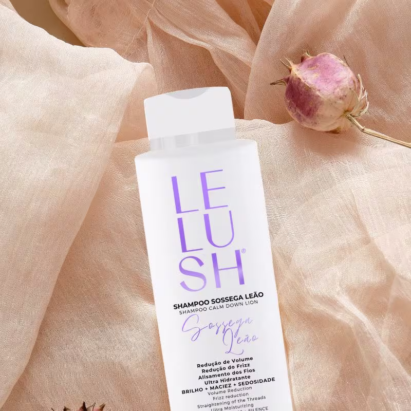 A imagem mostra um frasco de shampoo branco com o nome da linha escrito em letras roxas. A composição também inclui um tecido bege delicado e um botão de rosa rosada, criando uma atmosfera suave e natural.