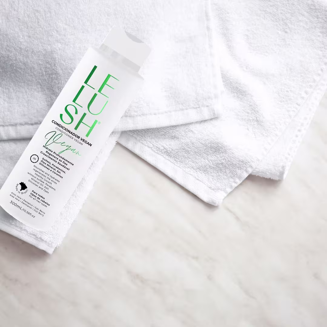 A imagem mostra o condicionador no canto da imagem. Ao lado da embalagem, há algumas toalhas brancas felpudas dobradas. A cena está em um ambiente claro, possivelmente um banheiro ou similar, com uma superfície aparentemente de mármore embaixo.