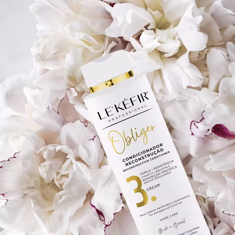 A imagem mostra o frasco do produto em destaque, rodeado por pétalas brancas de uma flor desmembrada. As pétalas têm algumas manchas avermelhadas. O cenário cria uma ambiente leve e delicado.