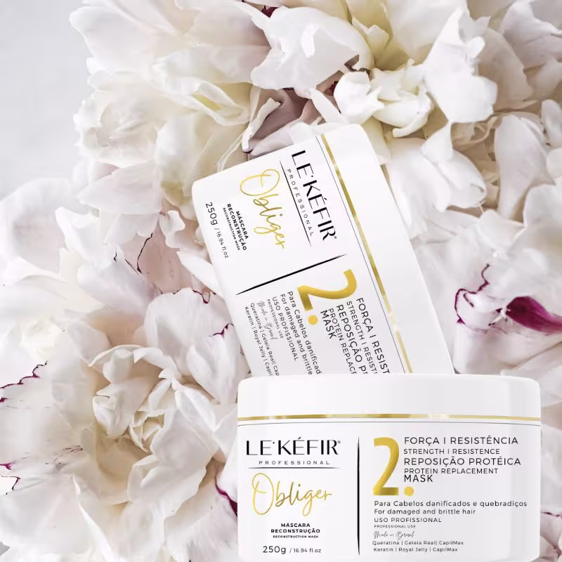 A imagem mostra duas embalagens da máscara capilar em destaque, rodeadas por pétalas brancas de uma flor desmembrada. As pétalas têm algumas manchas avermelhadas. O cenário cria uma ambiente leve e delicado.