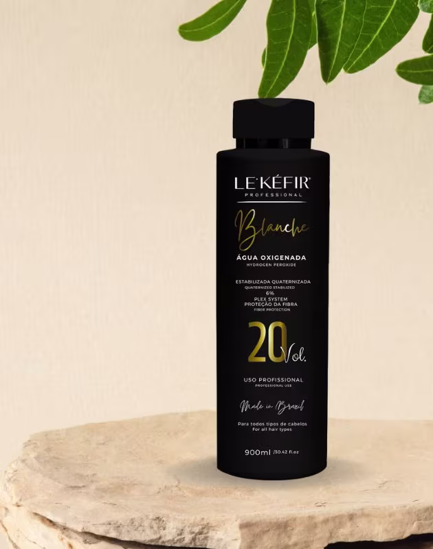 A imagem mostra a embalagem preta da água oxigenada posicionada sobre uma pedra clara, em um fundo bege, com folhas verdes decorando a cena, transmitindo uma atmosfera natural para esse cosmético capilar.