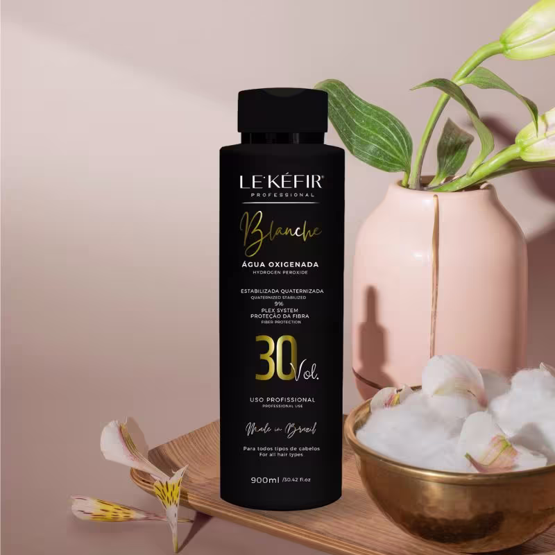 A imagem mostra a embalagem da água oxigenada posicionada ao lado de uma vasilha dourada com plantas e flores brancas, criando um cenário natural e elegante para apresentar o produto. As flores parecem ser orquídeas, com suas folhas verdes e hastes.