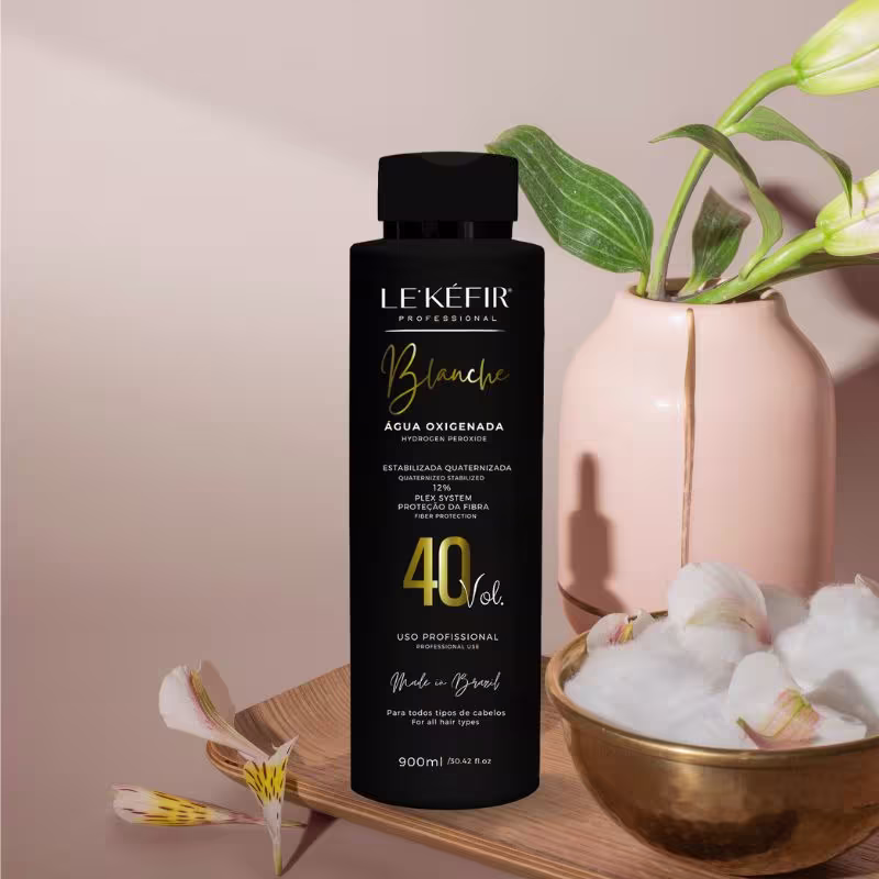 A imagem mostra a embalagem da água oxigenada posicionada ao lado de uma vasilha dourada com plantas e flores brancas, criando um cenário natural e elegante para apresentar o produto. As flores parecem ser orquídeas, com suas folhas verdes e hastes.