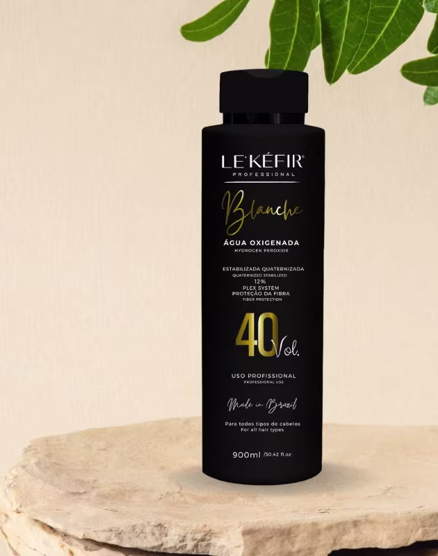 A imagem mostra a embalagem preta da água oxigenada posicionada sobre uma pedra clara, em um fundo bege, com folhas verdes decorando a cena, transmitindo uma atmosfera natural para esse cosmético capilar.