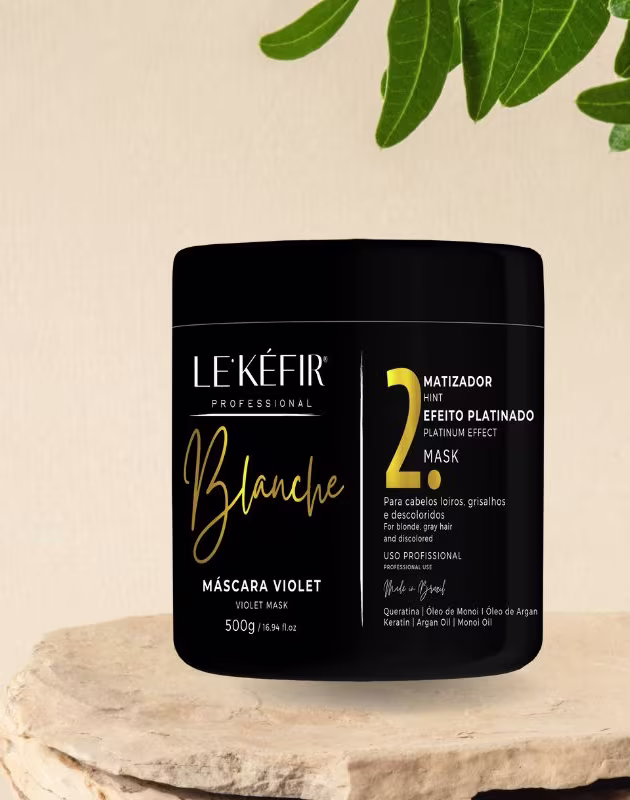 A imagem mostra a embalagem preta da máscara capilar posicionada sobre uma pedra clara, em um fundo bege, com folhas verdes decorando a cena, transmitindo uma atmosfera natural para esse cosmético capilar.