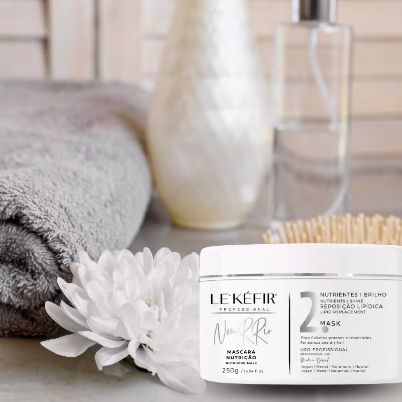 No primeiro plano, há o pote da máscara capilar "Le'Kéfir". 
Ao lado, há uma flor branca de crisântemo, adicionando um toque natural e delicado à composição. Nos fundos, vemos uma toalha felpuda cinza enrolada, um vidro transparente e outro frasco maior.