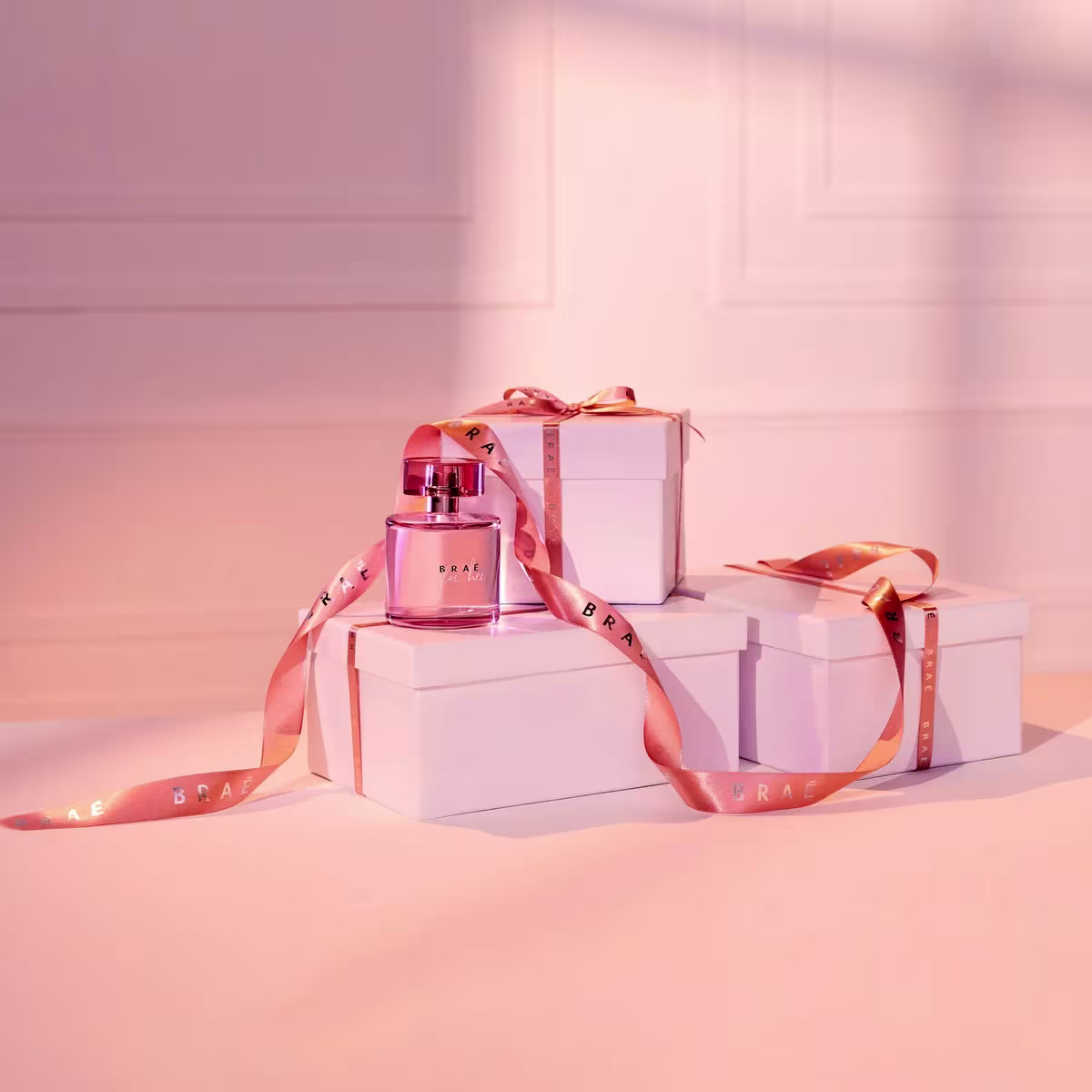 Perfume Braé em frasco de vidro rosa com tampa cromada. Caixas brancas retangulares com a marca Braé impressa em fitas rosa metálicas que as envolvem. As cores predominantes são rosa claro, branco e detalhes em rosa metálico.