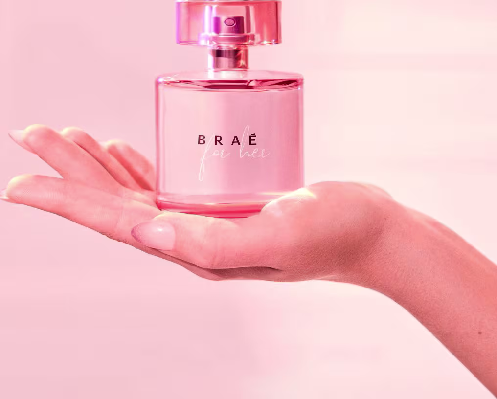 Perfume Braé em frasco redondo de vidro transparente rosa. O frasco tem uma tampa rosa brilhante. O perfume é segurado por uma mão feminina. O fundo é rosa claro, criando um ambiente suave e delicado para apresentar o produto de fragrância.