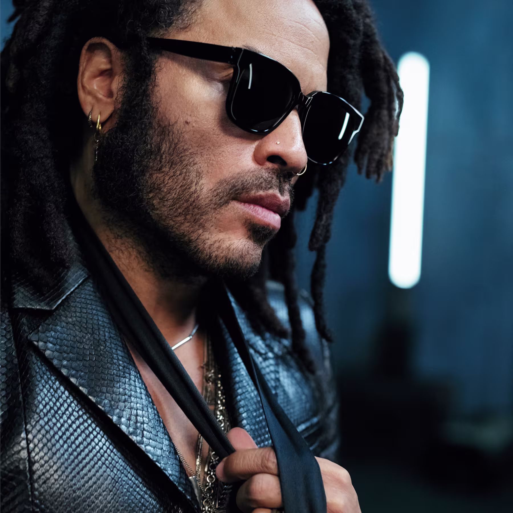 Homem de pele escura usando óculos escuros de armação preta, brincos dourados e um casaco azul escamado ou com textura de couro de réptil. Ele tem dreadlocks e uma barba curta e aparenta estar em um ambiente com iluminação dramática de fundo azul.