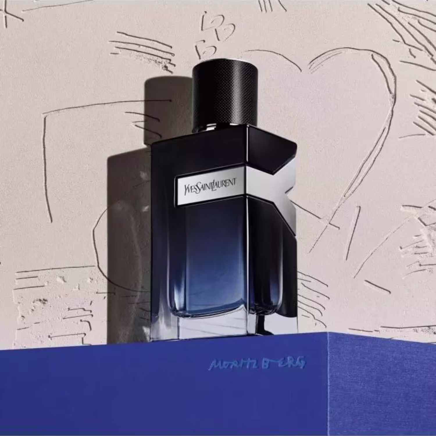Perfume em frasco retangular preto com detalhes metálicos na parte superior. O frasco é de vidro azul escuro translúcido. Na frente, está impresso o nome da marca "Yves Saint Laurent" em letras brancas. O frasco repousa sobre uma superfície azul.