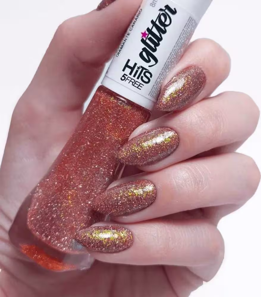 Esmalte de unhas líquido com brilho holográfico em tons de vermelho, laranja e dourado. Frasco de vidro transparente com tampa branca. Rótulo branco com o nome da marca "Hits" e a inscrição "5FREE" em letras pretas e rosa.