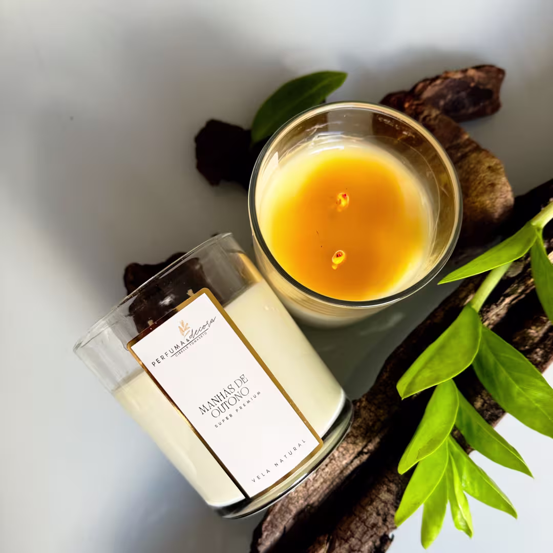 Vela em frasco de vidro transparente. Embalagem retangular branca com rótulo "Perfuma & Decora - Manhãs de Outono" em letras marrons. Ao lado, folhas verdes e pedaços de casca de árvore marrom dispõem-se sobre superfície clara.