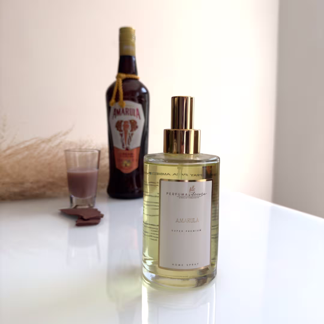 Home spray em frasco de vidro transparente. Embalagem cilíndrica dourada com rótulo branco indicando "Perfuma Decora Amarula". Garrafa de licor Marula com rótulo vermelho e elefante estampado ao lado. Copo com bebida marrom e pedaços de chocolate ao lado.