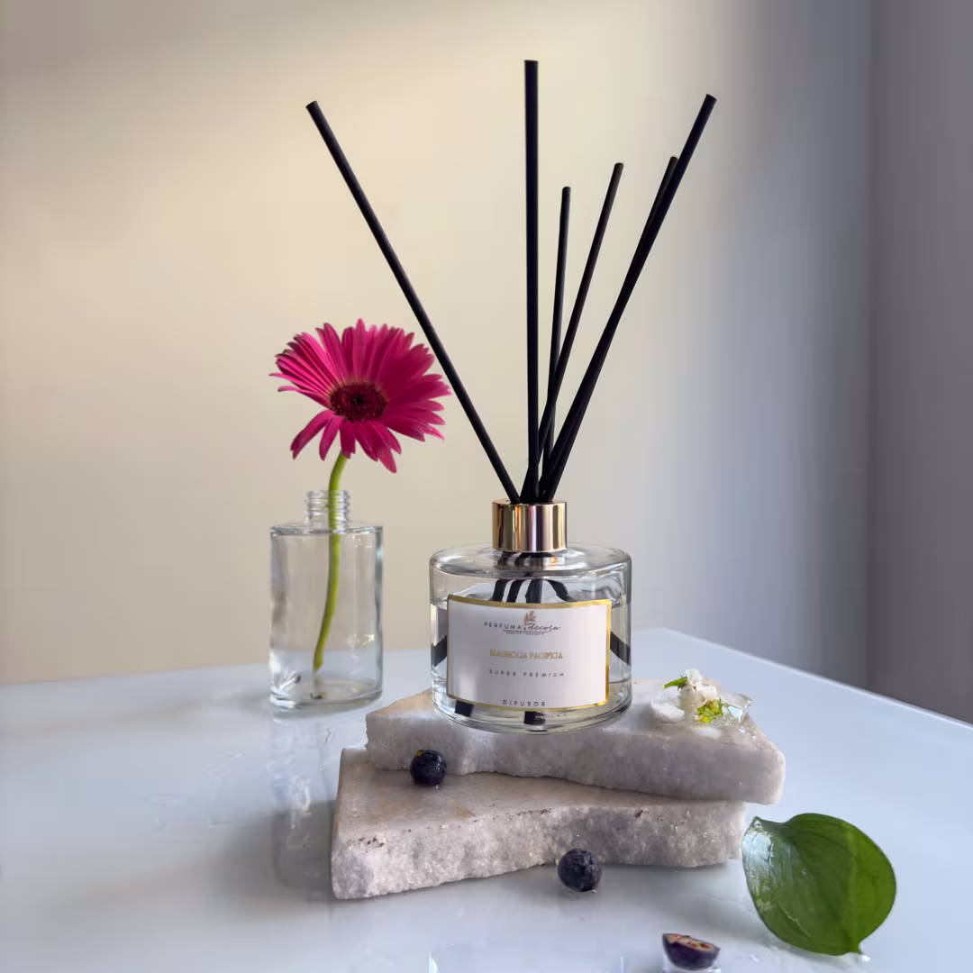 Frasco de vidro transparente com difusor de aromas em hastes pretas e frasco menor com uma gerbera rosa viva. Pedra porosa cinza com algumas amoras pretas e folha verde ao lado. Cores neutras com detalhes rosa vibrante da flor.