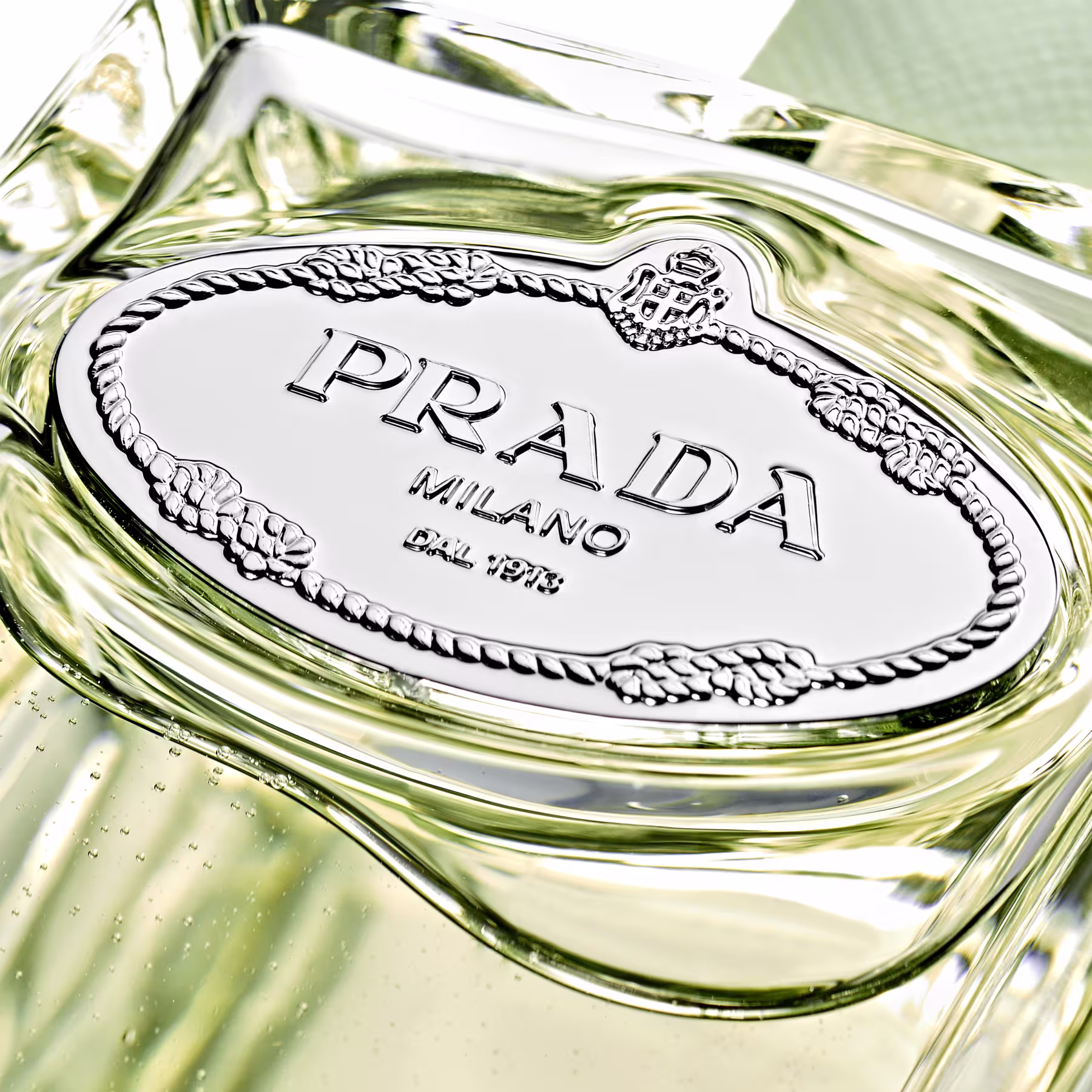 Perfume em frasco de vidro transparente com detalhes prateados. O frasco tem formato retangular com cantos arredondados. O rótulo oval prateado no corpo do frasco exibe o nome "PRADA" em letras maiúsculas, seguido de "MILANO" e o número "DAL 1913".