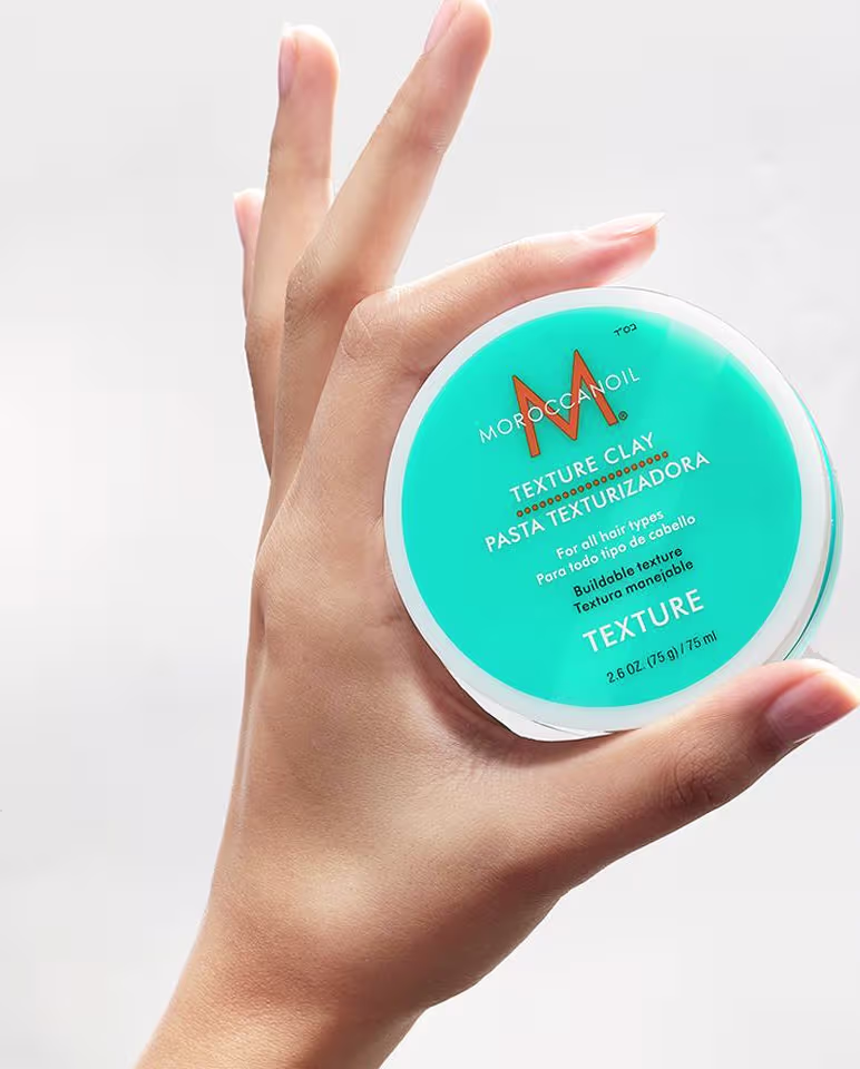 A imagem mostra uma mão feminina segurando o produto Moroccanoil Texture Clay, embalagem redonda de plástico na cor turquesa, com M laranja e texto com letras brancas e pretas. O fundo é neutro para destacar a imagem.