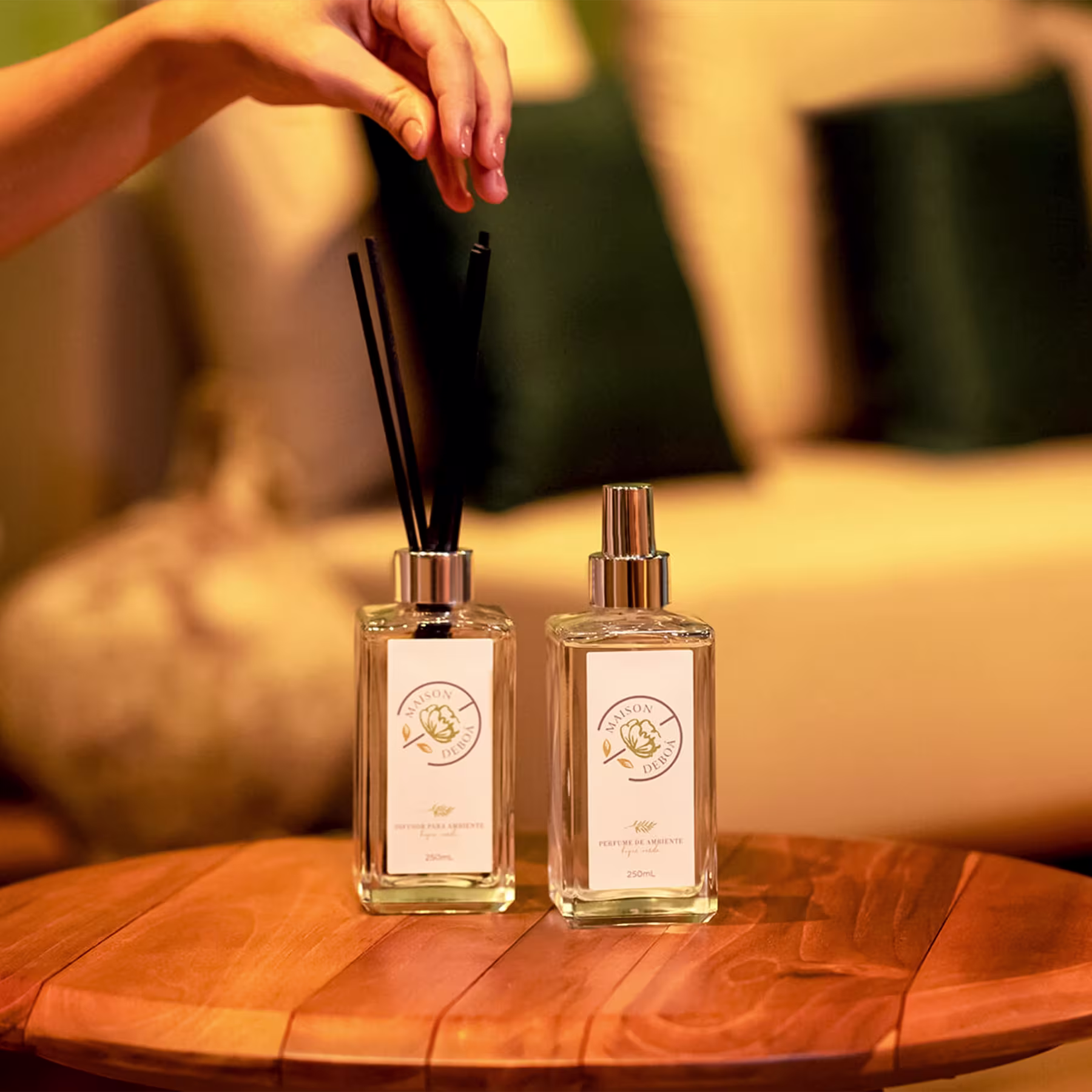 Dois frascos da marca Maison Deboá. Os frascos são de vidro transparente com detalhes dourados nas tampas e rótulos. Um dos frascos é de difusor de ambiente e outro um perfume de ambiente.