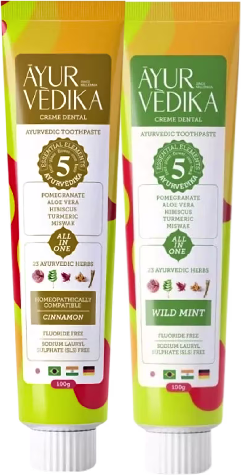 Dois tubos de creme dental, um com sabor de canela e outro de menta selvagem. Ambos os tubos possuem rótulos verdes e amarelos, com informações sobre os ingredientes Ayurvédicos utilizados, como romã, aloe vera, hibisco e curcuma.