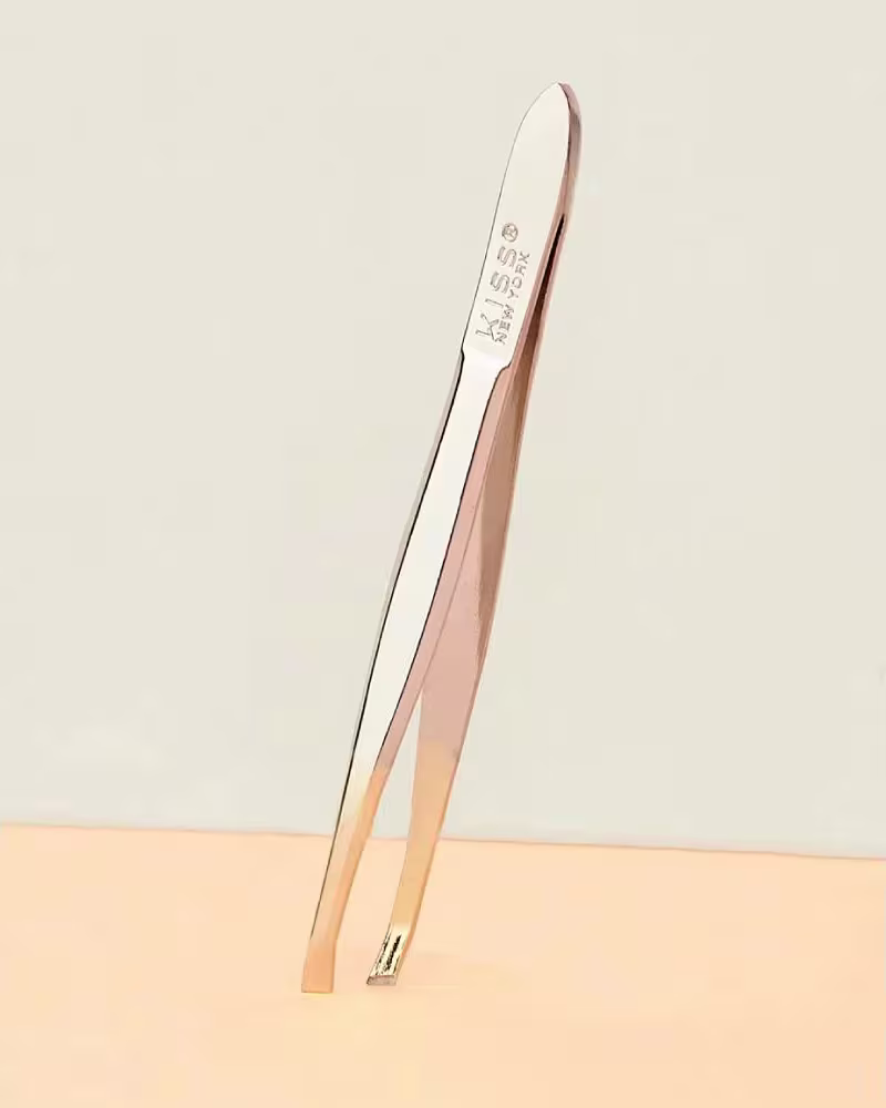 A imagem mostra a pinça de sobrancelha em metal com cabo longo e curvado. A pinça é de cor prateada com detalhes em dourado metálico.