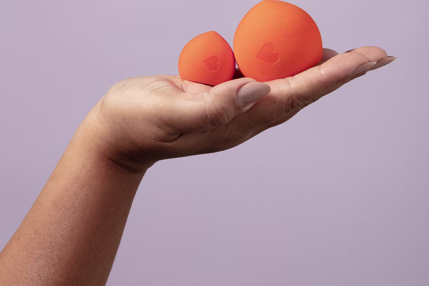 A imagem mostra duas esponjas de cor laranja, que parecem ser produtos de beleza da marca Quem Disse, Berenice?. Ambos estão sendo sendo segurados por uma mão, com um fundo liso de cor lilás.