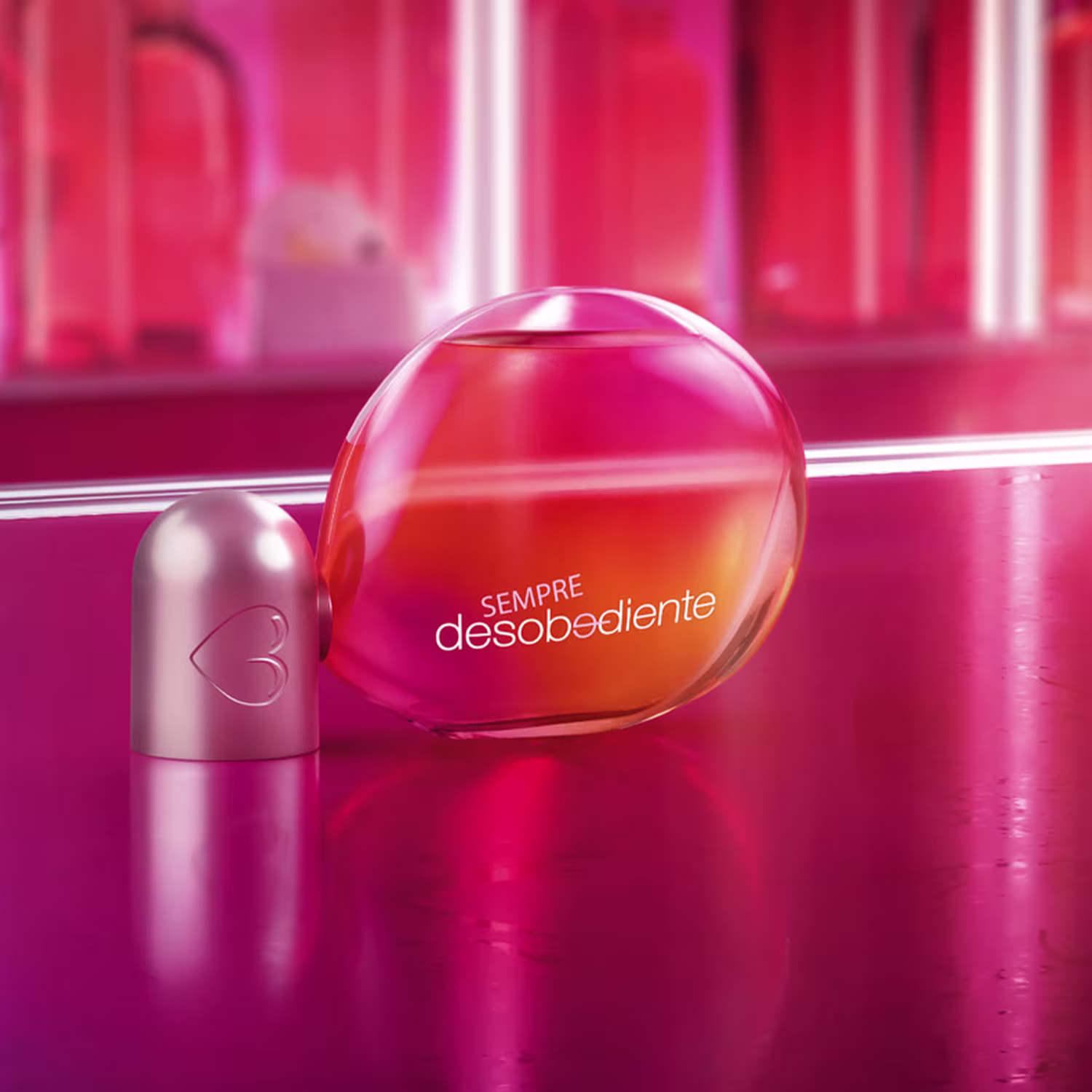 Fragrância em frasco esférico transparente com líquido alaranjado e texto "sempre desobediente" impresso. Embalagem em tons de rosa vibrante, com detalhes brilhantes e reflexos. Design minimalista e moderno, com foco no formato arredondado.