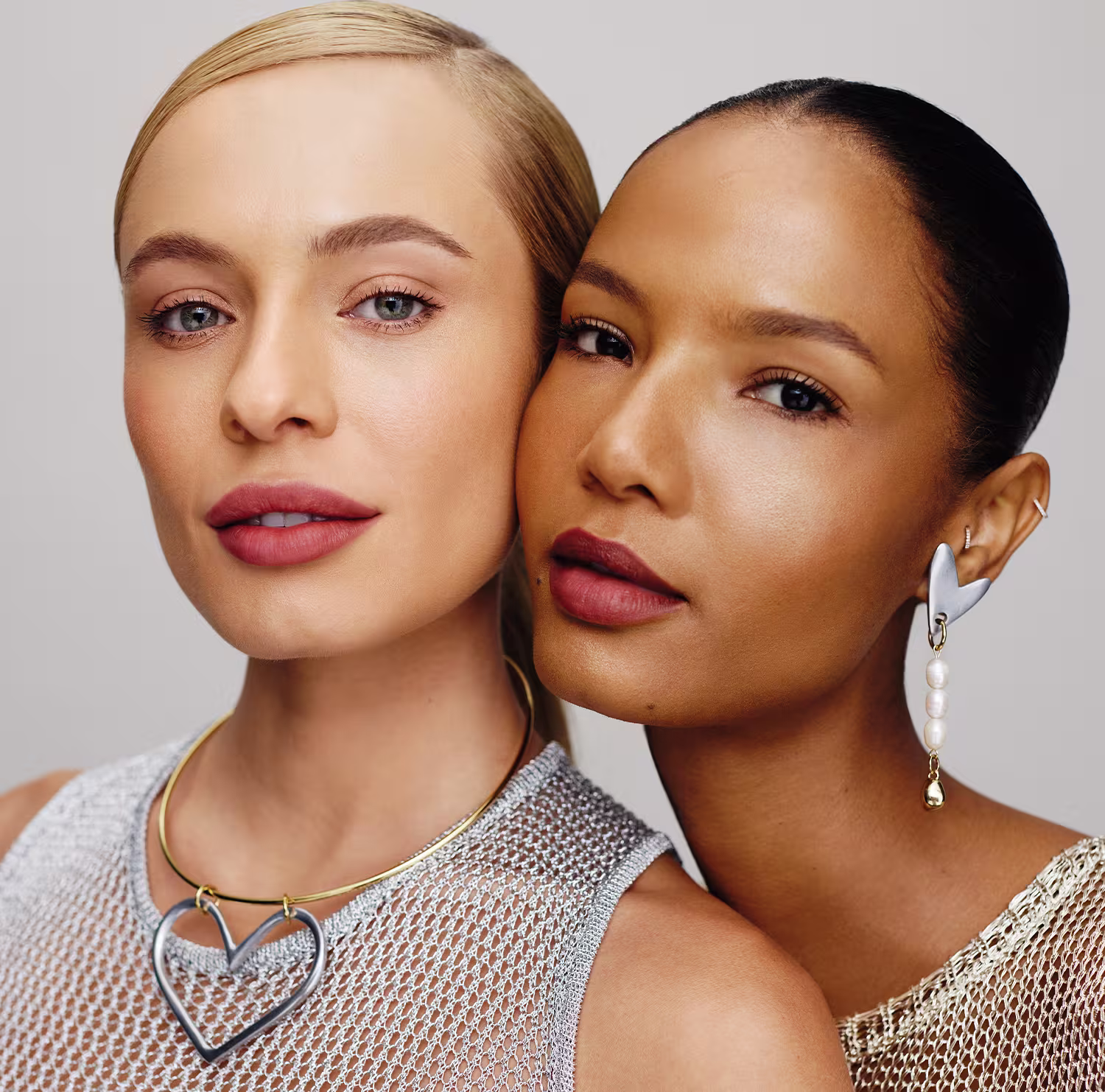 A imagem mostra dois rostos femininos em close-up, com expressões sérias e olhares diretos. Ambas as mulheres usam maquiagem com tons neutros e naturais, destacando seus traços faciais. Elas vestem roupas com detalhes em malha metálica.