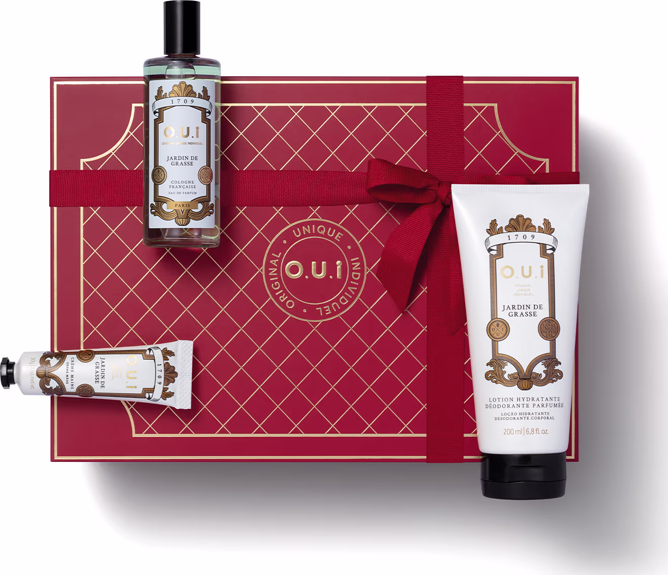 A imagem mostra uma bela caixa de presente vermelha com detalhes dourados e um lindo laço. Em cima da caixa há um frasco de vidro com o eau de parfum, um pequeno tubo com hidratante para as mãos e outro tubo maior com loção corporal.