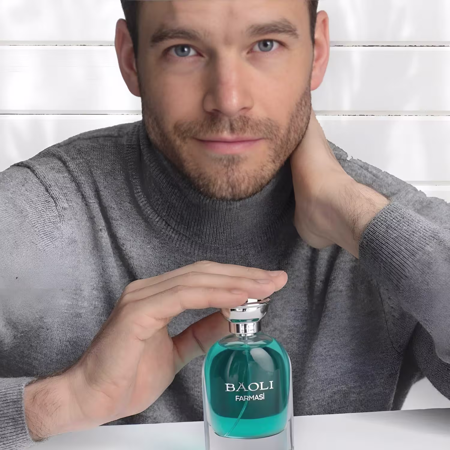 Perfume em frasco de vidro transparente com líquido de cor verde esmeralda. A tampa do frasco é prateada e brilhante. Um homem segura o frasco com uma das mãos, vestindo um suéter cinza de gola alta.