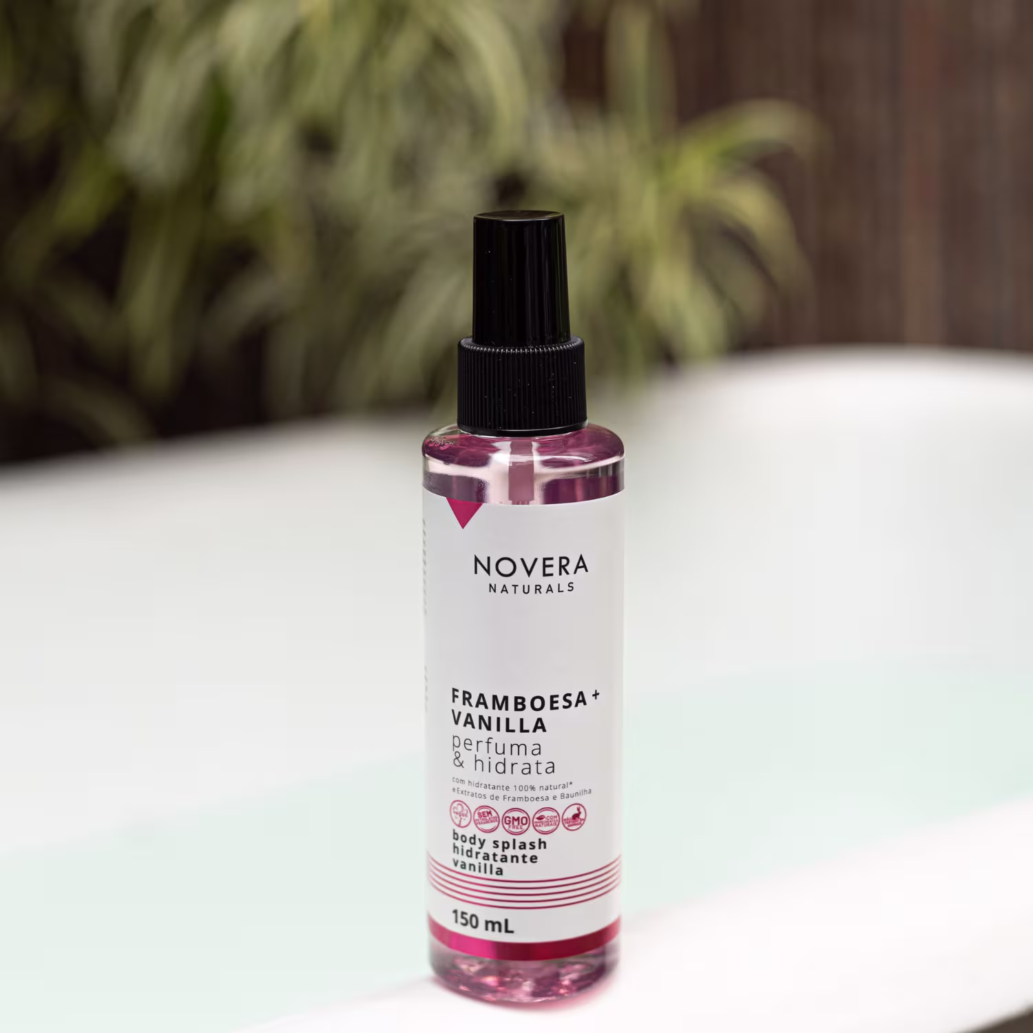 A imagem mostra o body splash Novera Naturals Framboesa + Vanilla. O frasco é de 150ml, transparente, permitindo ver o líquido rosa claro dentro. Ele está sobre uma superfície branca e há algumas plantas ao fundo.