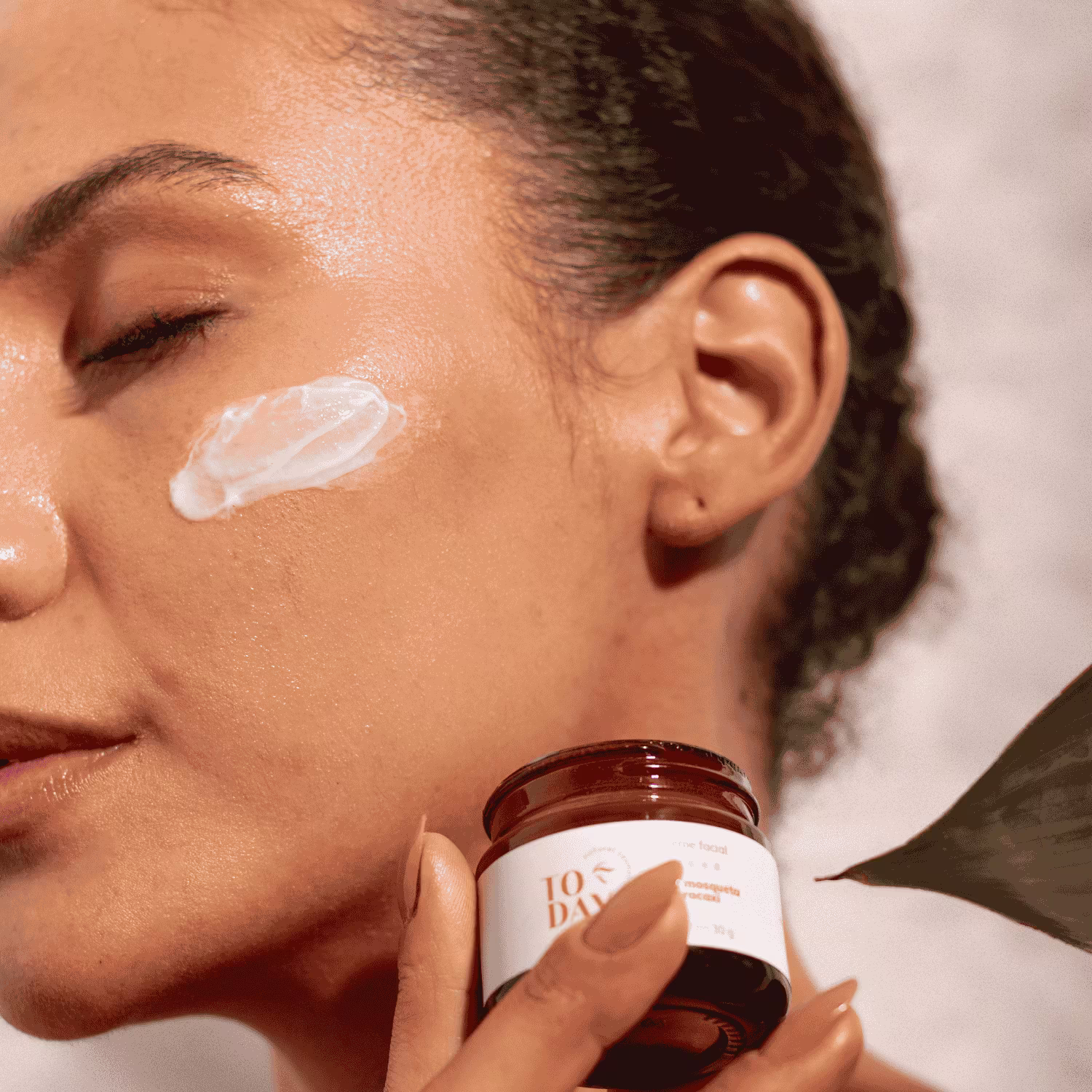 A imagem mostra o rosto de uma mulher morena com os olhos fechados e os cabelos presos, com um pouco de creme aplicado nas maçãs do rosto. Ela segura o pote do creme Rosa Mosqueta ao lado do rosto.