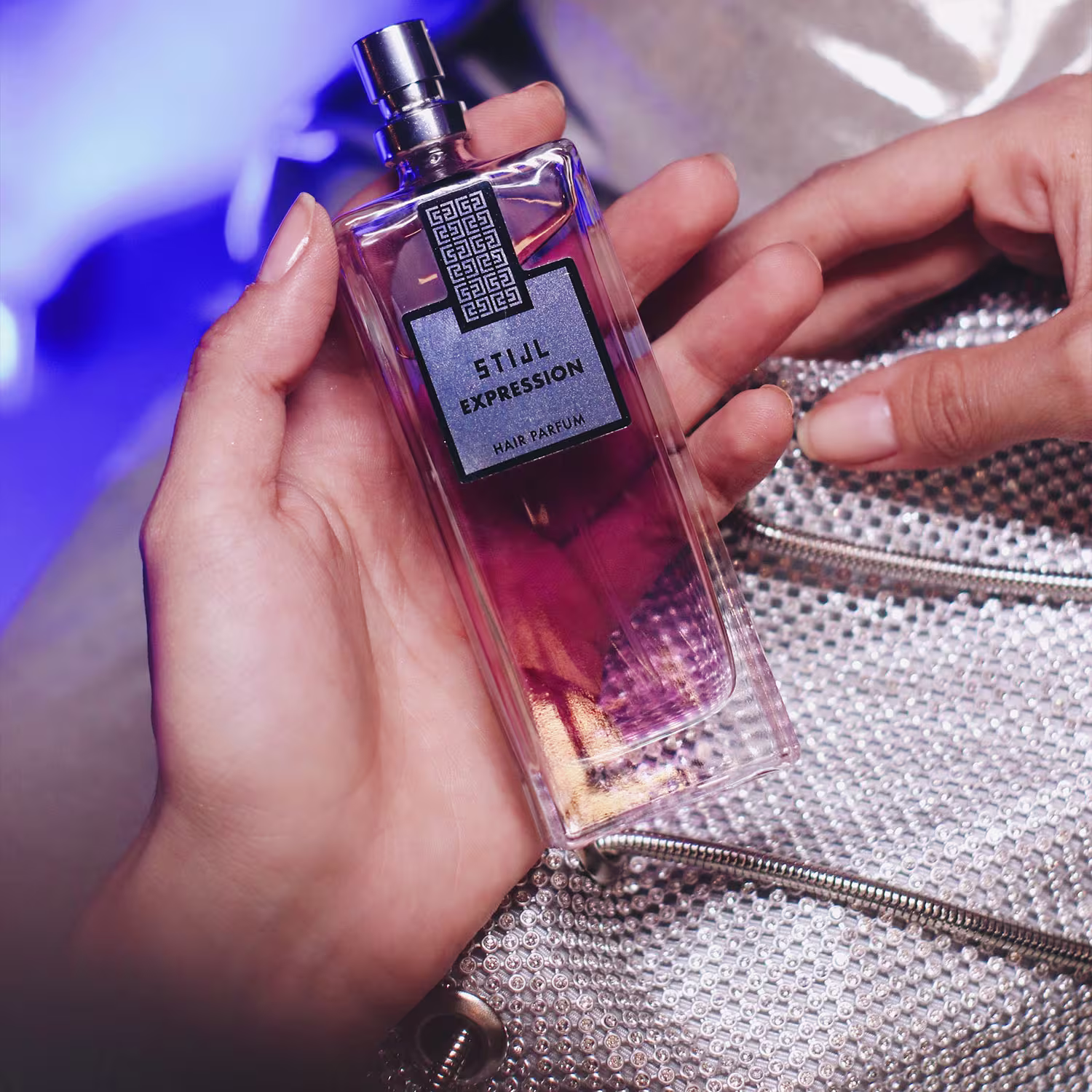 Perfume para cabelos em frasco retangular transparente contendo líquido rosa e dourado. O frasco tem uma tampa metálica. O rótulo preto e cinza exibe o texto "Stijl Expression Hair Parfum" com um padrão geométrico.